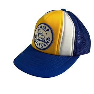 May include: A yellow, white, and blue trucker hat with a blue and white patch that says "Camp Whitesand" with an elk inside the circle.