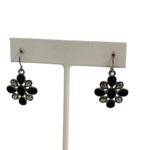 May include: A pair of silver-toned drop earrings. Each earring features a floral design with black and silver-colored stones. The earrings are displayed on a white earring stand against a white background.