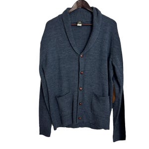 May include: A dark blue cardigan with a shawl collar, five brown buttons, and two patch pockets. The cardigan has a brown leather patch on the right elbow.