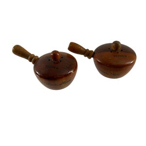 May include: A set of two wooden salt and pepper shakers with a brown wood finish. The shakers are shaped like small pots with handles and have the words "Salt" and "Pepper" written on them.