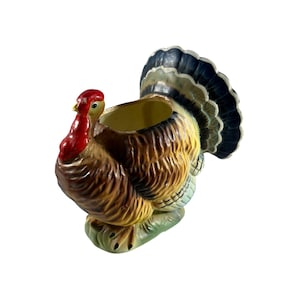 May include: A ceramic turkey-shaped planter with a red head and wattle, brown body, and a fan of blue, yellow, and white tail feathers. The planter is perched on a green base, perfect for seasonal decor.