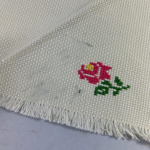May include: A white fabric with a fringed edge and a pink and green embroidered rose.