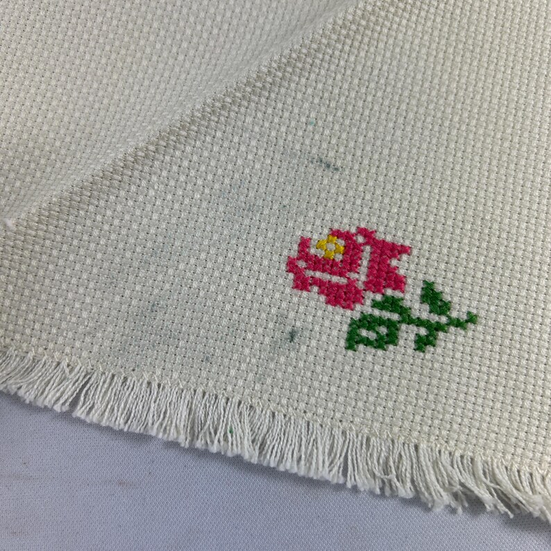 May include: A white fabric with a fringed edge and a pink and green embroidered rose.