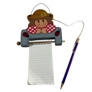 May include: A wooden notepad holder featuring a painted girl with a straw hat and red checkered sleeves. The notepad has lined paper and a purple pencil attached with string. The holder is painted blue with ladybug details.