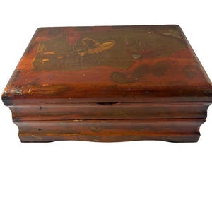 May include: A rectangular wooden box with a hinged lid. The box is stained a reddish-brown color with a painted design on the lid featuring gold and brown accents. The box has a decorative base.