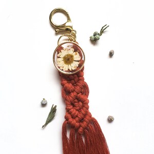 May include: A red macrame keychain with a small round pendant featuring a pressed white daisy. The keychain has a gold metal clasp and a long tassel.