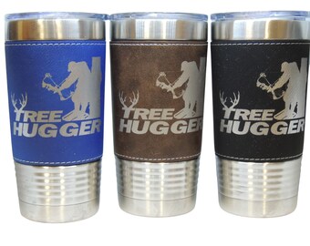 Puede incluir: Tres vasos de acero inoxidable con envoltura de cuero. Los vasos son azul, marrón y negro. Cada vaso tiene una silueta de una persona sosteniendo un arco y una flecha y el texto "TREE HUGGER".