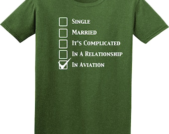 May include: A dark olive green short-sleeved t-shirt featuring a white graphic of a checklist with the text Single, Married, It's Complicated, In A Relationship, and In Aviation, with a checkmark placed next to the In Aviation option.
