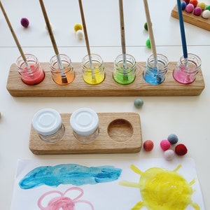Op de afbeelding: Een houten verfhouder met zes kleine glazen potjes gevuld met verschillende kleuren verf. De potjes hebben houten deksels en elk heeft een penseel in de verf gestoken. Er zijn twee extra kleine glazen potjes met witte deksels op een aparte houten lade. Een wit vel papier met een geverfde bloem, zon en gras is op de achtergrond.