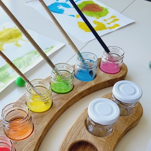 Puede incluir: Una paleta de pintura de madera con siete pequeños frascos de vidrio llenos de pintura colorida. La paleta tiene forma de media luna y tiene un compartimento separado para pinceles. Los colores de la pintura son rojo, naranja, amarillo, verde, azul y rosa.