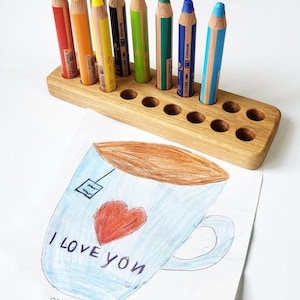 May include: A wooden pencil holder with ten holes, holding a variety of colored pencils. The pencils are red, orange, yellow, brown, black, green, blue, and light blue. A drawing of a teacup with the words "I LOVE YOU" is in the foreground.