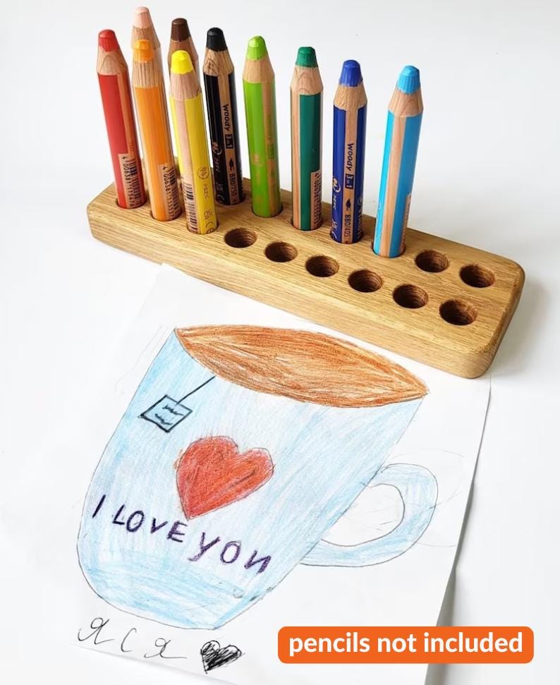 May include: A wooden pencil holder with ten holes, holding a variety of colored pencils. The pencils are red, orange, yellow, brown, black, green, blue, and light blue. A drawing of a teacup with the words "I LOVE YOU" is in the foreground.