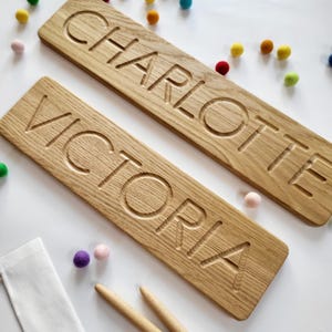 Peut inclure: Deux planches rectangulaires en bois clair avec les prénoms CHARLOTTE et VICTORIA gravés en lettres capitales. Les planches sont entourées de petites boules en feutre colorées et de deux stylets de traçage en bois sur un fond blanc.