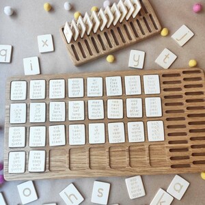 May include: Wooden educational toy with letter and word tiles. The set includes a wooden tray with slots for the tiles, individual letter tiles, and word tiles with Dutch words. The tiles are off-white with black lettering. Several small, colorful pom-poms are scattered around the tray.
