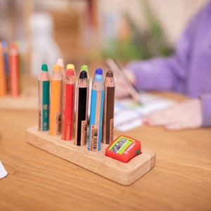 May include: A set of colorful pencils in a wooden holder with a red pencil sharpener. The pencils are arranged in a row, with a variety of colors including green, orange, red, black, blue, and brown. The pencils are on a wooden table.