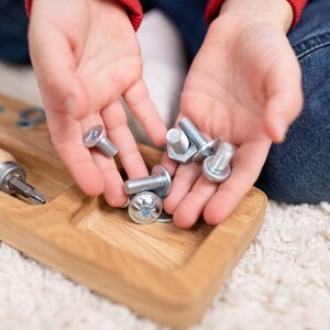 May include: A child's hands holding a variety of silver screws and nuts. The screws and nuts are on a wooden tray with a screwdriver.