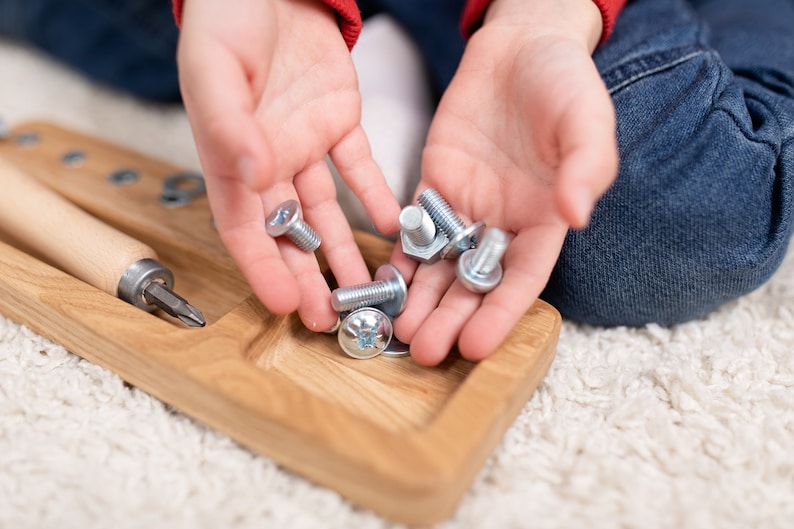 May include: A child's hands holding a variety of silver screws and nuts. The screws and nuts are on a wooden tray with a screwdriver.