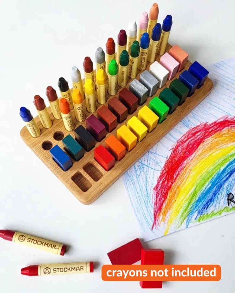 May include: Wooden crayon organizer with a variety of colorful crayons and crayon blocks. The organizer is made of light-colored wood and has several slots to hold the crayons. A drawing of a rainbow is in the background. The text "crayons not included" is at the bottom.