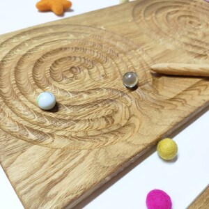 May include: Wooden board with two spiral grooves for marble play. The board is made of light brown wood and has a natural finish. Two marbles are in the grooves, one white and one clear. There are also several small, colorful felt balls scattered around the board.
