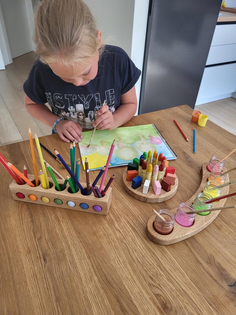 Puede incluir: Un portal&aacute;pices rectangular de madera con incrustaciones circulares de colores, un bloque de madera circular que sostiene crayones en posici&oacute;n vertical y una bandeja de madera curva con cinco frascos de vidrio peque&ntilde;os llenos de pintura l&iacute;quida.