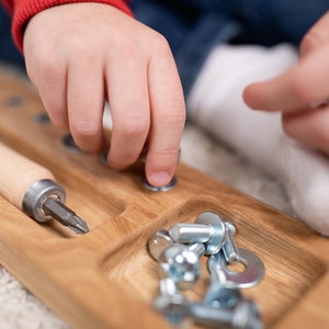 May include: A wooden tray with a screwdriver and a compartment filled with screws and nuts. A child's hand is shown placing a nut on a screw.