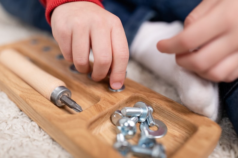 May include: A wooden tray with a screwdriver and a compartment filled with screws and nuts. A child's hand is shown placing a nut on a screw.