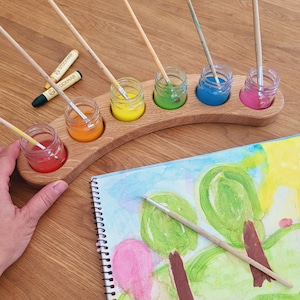 May include: A wooden paint palette with six small glass jars filled with different coloured paints. The jars are arranged in a semi-circle and each has a wooden paintbrush. A sketchbook with a watercolour painting of trees is on the table.