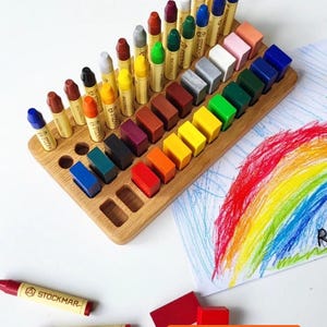 Könnte beinhalten: Ein hölzerner Buntstifthalter mit einer Vielzahl von farbigen Buntstiften und Wachsblöcken. Der Halter ist mit verschiedenfarbigen Buntstiften und Wachsblöcken gefüllt. Rechts ist eine Zeichnung eines Regenbogens. Der Text "crayons not included" steht unten.