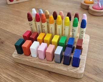May include: A wooden tray with 16 compartments holds a set of 24 colorful crayons. The crayons are arranged in rows and are labeled "Wachsmalstifte" and "Stoffmalfarben".