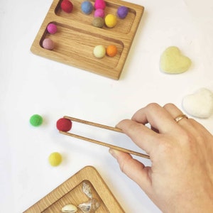 Puede incluir: Una bandeja de madera con dos compartimentos, cada uno lleno de bolas de fieltro de colores. Una mano usa palillos de madera para recoger una bola de fieltro roja. Tambi&eacute;n hay dos corazones de fieltro en el fondo.