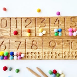 May include: Wooden number tracing board with numbers 0-10 carved into the surface. Colorful felt balls are placed next to each number. Two wooden sticks are at the bottom. The board is made of light brown wood.