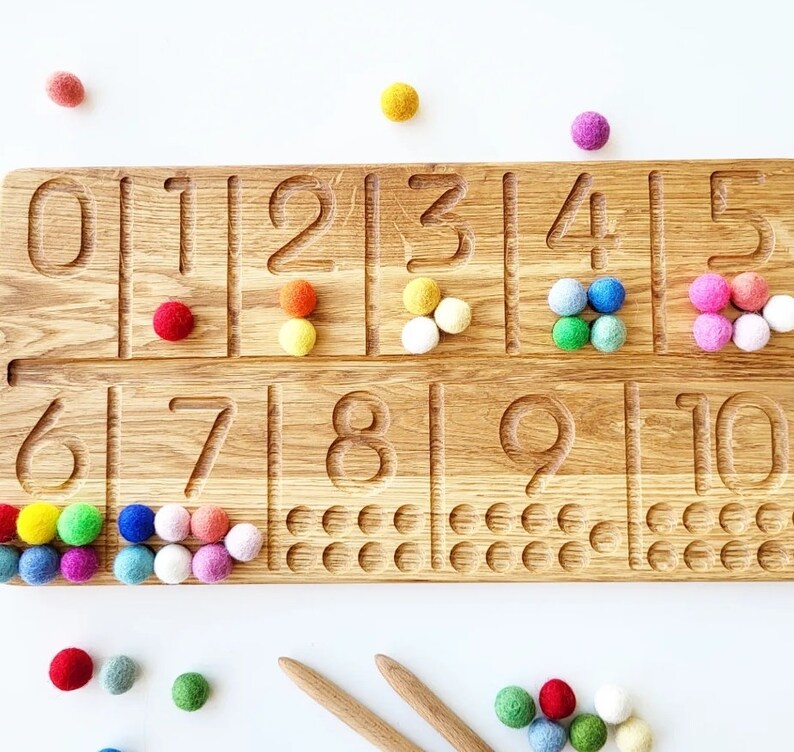 May include: Wooden number tracing board with numbers 0-10 carved into the surface. Colorful felt balls are placed next to each number. Two wooden sticks are at the bottom. The board is made of light brown wood.
