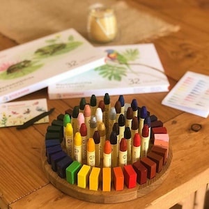 May include: A set of 32 colorful crayons arranged in a circle on a wooden tray. The crayons are labeled "STOCKMAR WACHSMALSTIFT" and are arranged by color, with the darkest colors on the outside and the lightest colors on the inside.