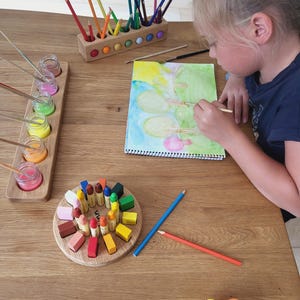 Puede incluir: Un portal&aacute;pices de madera con l&aacute;pices de colores, un soporte de madera con frascos de vidrio de pintura, un bloque circular de madera con ceras de colores y un cuaderno de bocetos en espiral con una pintura de acuarela de &aacute;rboles.