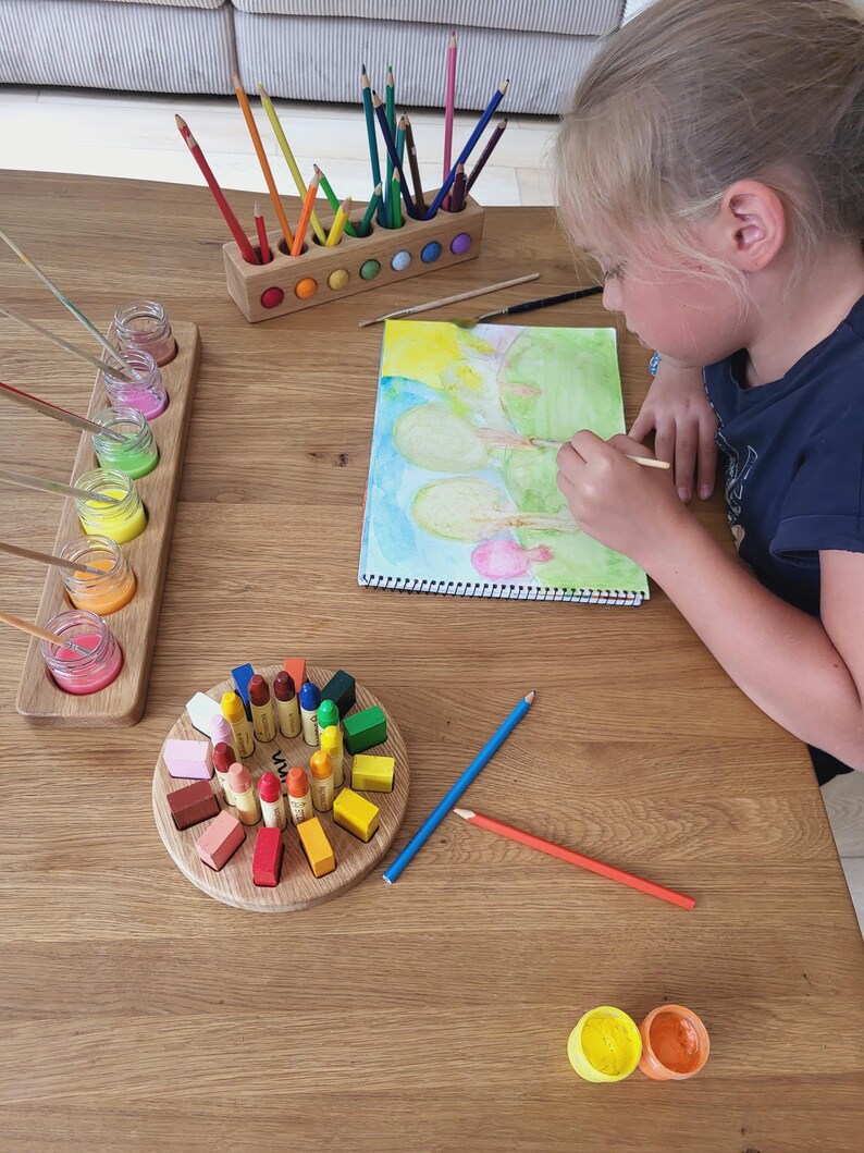 Puede incluir: Un portal&aacute;pices de madera con l&aacute;pices de colores, un soporte de madera con frascos de vidrio de pintura, un bloque circular de madera con ceras de colores y un cuaderno de bocetos en espiral con una pintura de acuarela de &aacute;rboles.