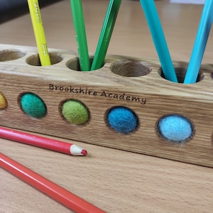 Puede incluir: Un portal&aacute;pices rectangular de madera con aberturas circulares que contienen l&aacute;pices de colores y bolas de fieltro. La madera est&aacute; grabada con el texto Brookshire Academy. L&aacute;pices rojos y naranjas yacen horizontalmente frente al soporte.