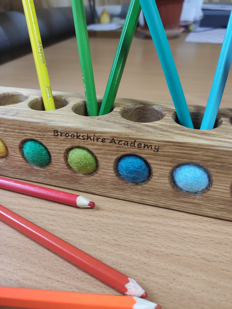 Puede incluir: Un portal&aacute;pices rectangular de madera con aberturas circulares que contienen l&aacute;pices de colores y bolas de fieltro. La madera est&aacute; grabada con el texto Brookshire Academy. L&aacute;pices rojos y naranjas yacen horizontalmente frente al soporte.