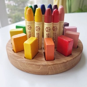 May include: A wooden round tray with a circular cutout for crayons. The tray has 12 slots for crayons, and the crayons are arranged in a circle around the tray. The crayons are all different colors, including red, orange, yellow, green, blue, and purple. The tray is made of natural wood and has a smooth finish.