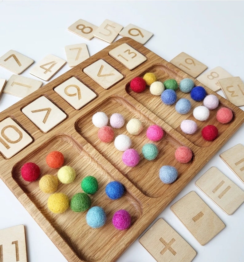 May include: Wooden number learning set with a divided tray and colorful felt balls. Includes wooden number cards and a tray with compartments for sorting. The felt balls are in various colors, including red, yellow, and blue.