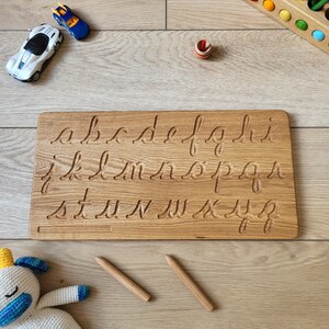 May include: Wooden lowercase alphabet tracing board with grooves for each letter. Two wooden pencils are included. A white and blue toy car, a blue and yellow toy car, a spinning top, and a wooden pencil holder with colored pencils are also visible.
