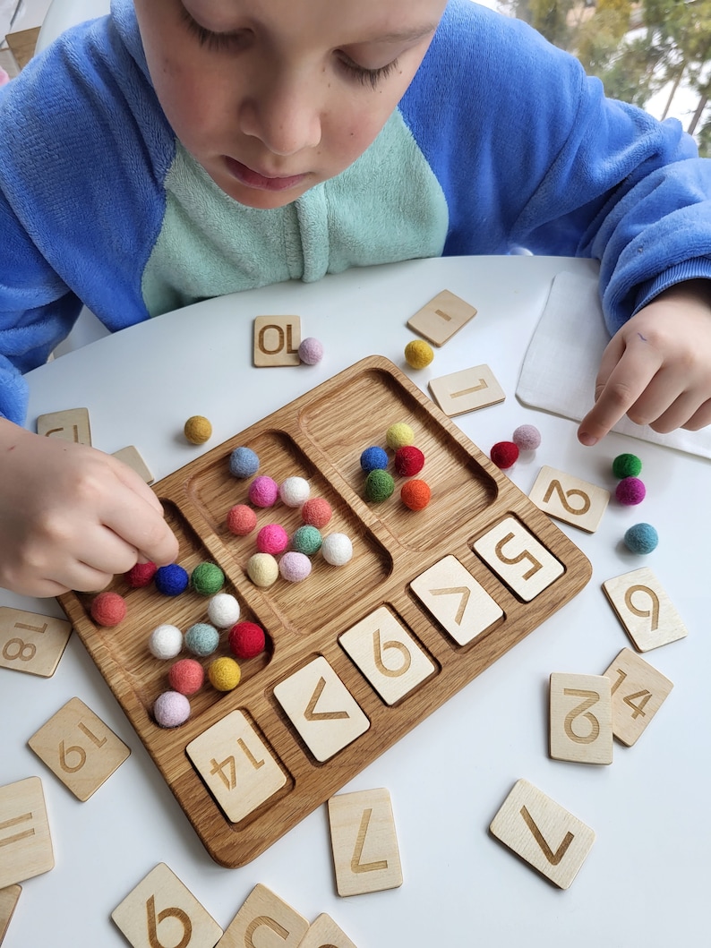 May include: Wooden educational toy with a tray, colorful felt balls, and wooden number tiles. The tray has compartments for sorting. Number tiles include the numbers 1, 2, 5, 6, 7, 9, 14, 18, and 19. The text "OL" is also visible.