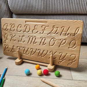 May include: Wooden alphabet tracing board with a stand, featuring uppercase cursive letters carved into the surface. The board is accompanied by colourful felt balls and wooden pencils.