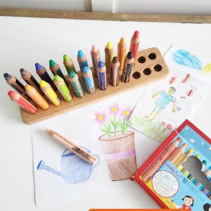 May include: A wooden pencil holder with a variety of colored pencils arranged in it. The holder has a rectangular base with angled slots for the pencils and several round holes. Drawings and a box of colored pencils are also visible.