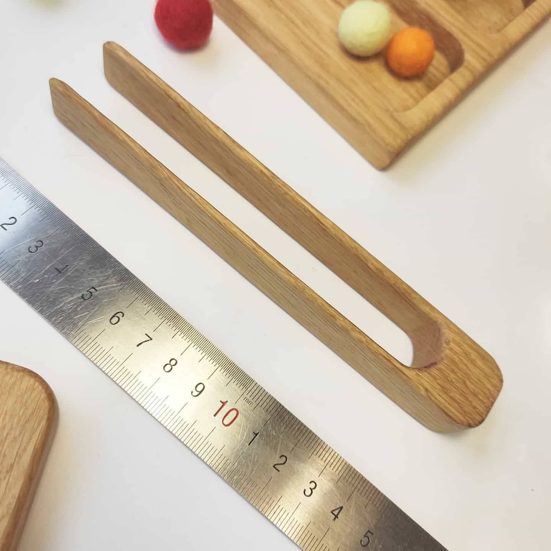Puede incluir: Pinzas de madera con un mango redondeado y una abertura rectangular. Las pinzas miden aproximadamente 25 cm de largo y est&aacute;n hechas de madera natural.