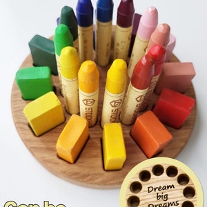 May include: A wooden crayon holder with a circular base and 12 slots for crayons. The holder is filled with colorful crayons and has a small wooden circle with the text "Dream big Dreams" on it.
