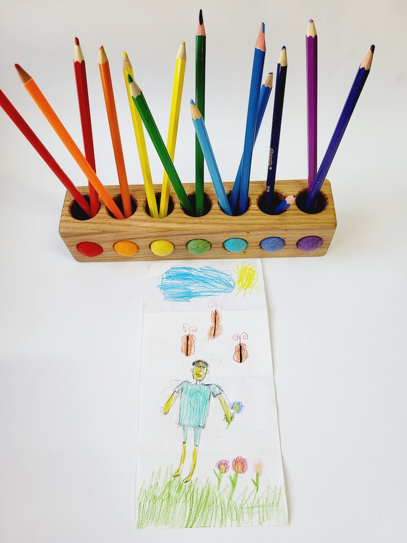 Puede incluir: Un portal&aacute;pices rectangular de madera con siete orificios circulares, cada uno con un l&aacute;piz de color. La parte frontal del soporte presenta siete inserciones circulares de fieltro en colores del arco&iacute;ris. Debajo hay un dibujo de una persona, flores y mariposas.