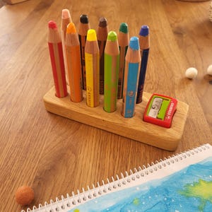 May include: A rectangular wooden pencil holder containing nine coloured pencils in shades of red, orange, yellow, green, blue, brown, and black, with a red Stabilo pencil sharpener embedded in a dedicated slot on the right side of the wooden base.
