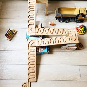Puede incluir: Una pista de canicas de madera de color claro con caminos serpenteantes tallados, que presenta una sección superior con canicas coloridas, una unión central y un final en espiral. La pista está ensamblada en segmentos sobre el suelo, junto a un camión volquete de juguete amarillo.