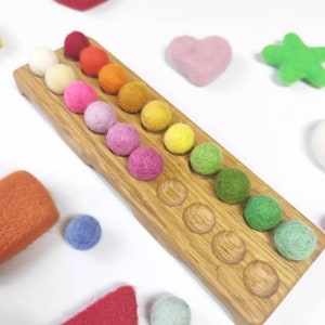 Könnte beinhalten: Ein Holztablett mit 18 bunten Filzkugeln in einem Regenbogen-Farbverlauf. Das Tablett hat 18 kreisrunde Vertiefungen für die Kugeln. Die Kugeln sind in einer Reihe von Rot nach Grün angeordnet.