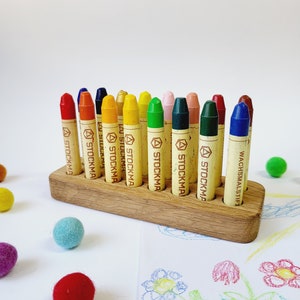 May include: A wooden crayon holder filled with colorful crayons. The crayons are arranged in a row and are various colors, including red, orange, yellow, green, and blue. The holder sits on a white surface with colorful felt balls scattered around.